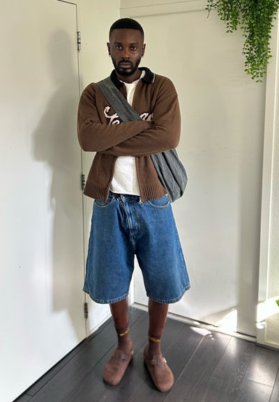 Cardigan marrón con texto bordado, camisa blanca, pantalones cortos de mezclilla azules, zuecos marrones y bandolera gris. Diseño sencillo y ajuste relajado.