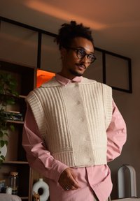 Gilet en tricot beige avec un texture côtelée, large conception d'épaules, porté sur une chemise à boutons rose clair. Les accessoires incluent des bagues et un matériel visible.