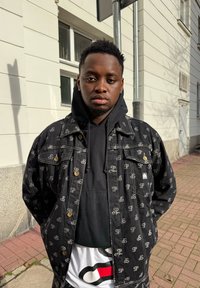 Casaco de ganga preto com padrão repetido de logótipo cinzento, usado por cima de um hoodie preto. O design inclui botões dourados e um ajuste folgado.