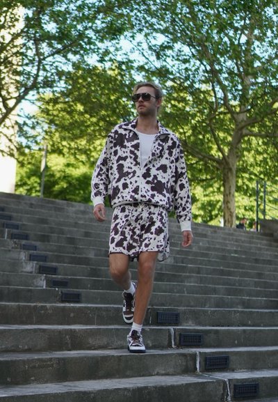 Conjunto de ropa activa de estampado de vaca en blanco y negro con chaqueta de cremallera y pantalones cortos. La modelo lleva gafas de sol y zapatillas deportivas. Fondo de escaleras urbanas.