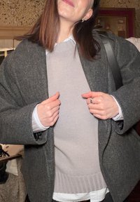Manteau gris foncé surdimensionné par-dessus un pull tricoté gris clair, superposé avec une chemise à rayures bleues et blanches. Bague visible sur la main gauche.