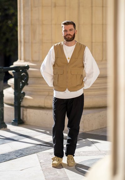 Chaleco beis con cremallera sobre un sudadera blanca, pantalones negros ajustados y zapatillas beis. Entorno natural con fondo arquitectónico.