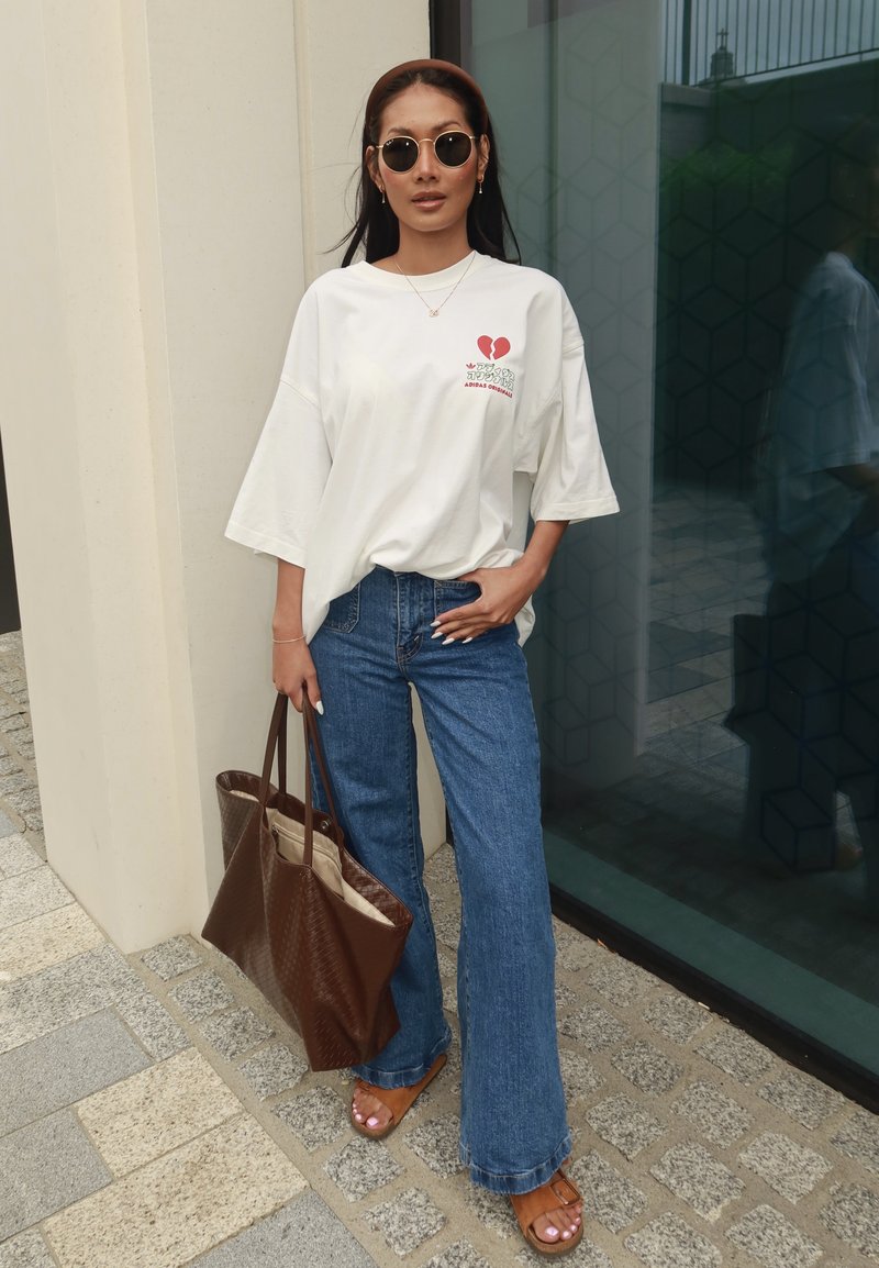 Femme portant des lunettes de soleil, un t-shirt blanc ample avec motif rouge, un jean bleu, des sandales brunes et un grand cabas brun, debout à l'extérieur.