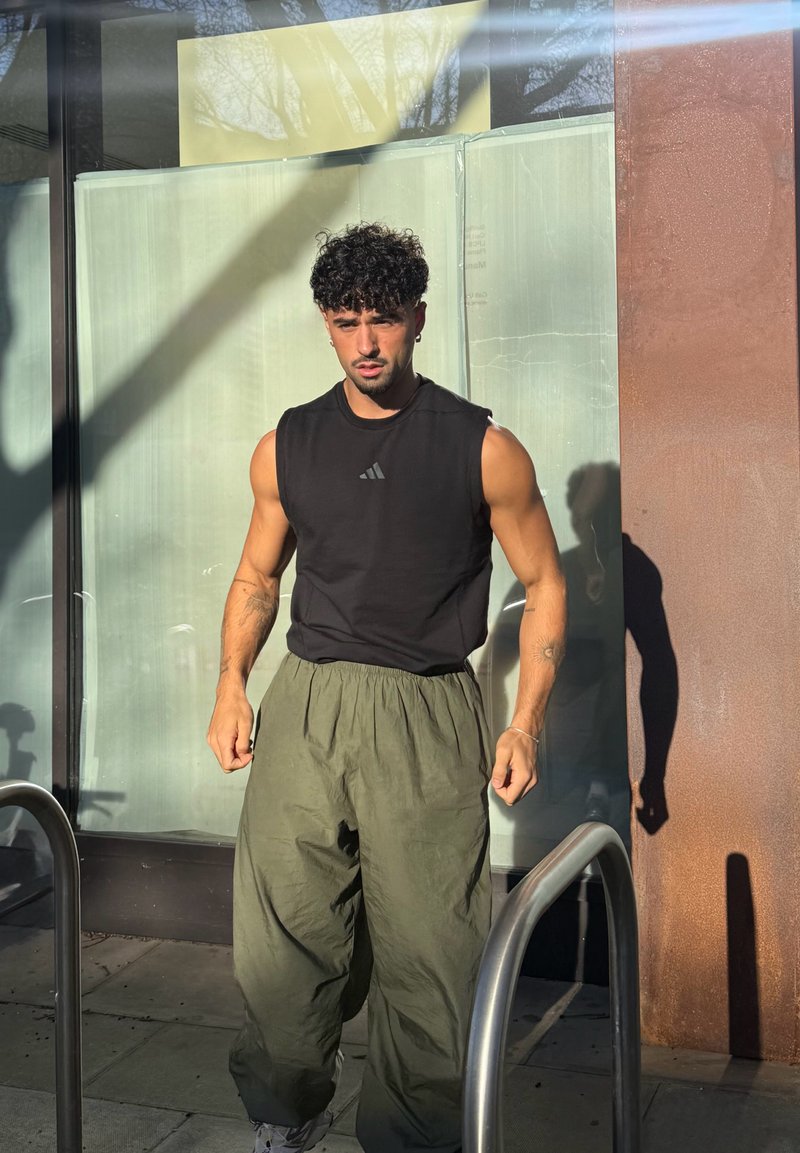 Haut noir sans manches avec logo gris, pantalon ample vert olive, debout devant un fond en verre, projetant une ombre au sol.