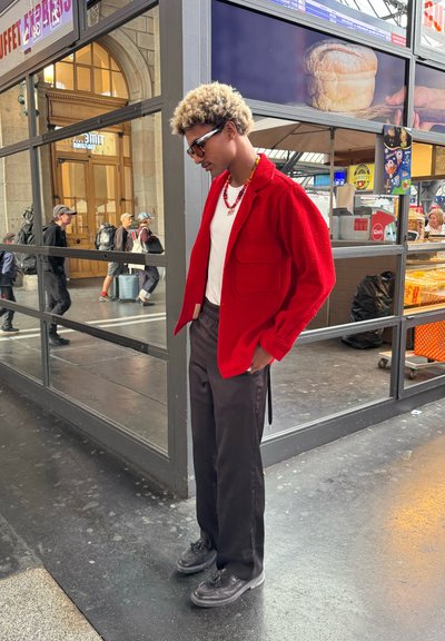 Persona joven con cabello rizado y rubio, que lleva una chaqueta roja, camisa blanca y pantalones negros, de pie cerca del mostrador de una panadería dentro de una estación concurrida.