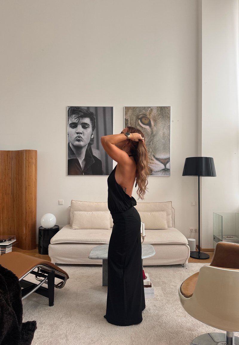Robe maxi noire sans manches avec un dos nu, texture lisse. La pièce présente des tons neutres, un canapé beige et une lampe noire.
