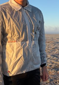 Giacca bianca leggera e stropicciata con chiusura a cerniera e collo abbassabile. Presenta grafiche verdi a contrasto e un piccolo logo rosso.