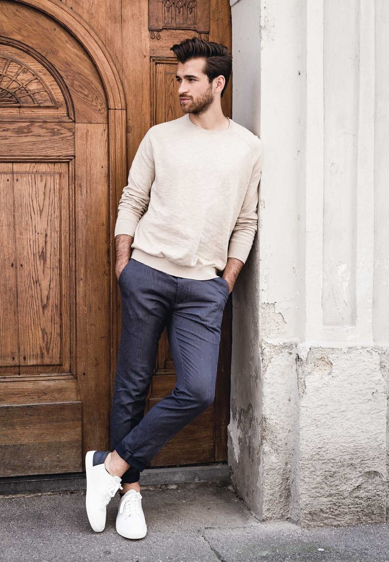 Beige trui, donkere broek met een subtiel patroon en witte sneakers. Staand tegen een gestructureerde muur en een houten deur.