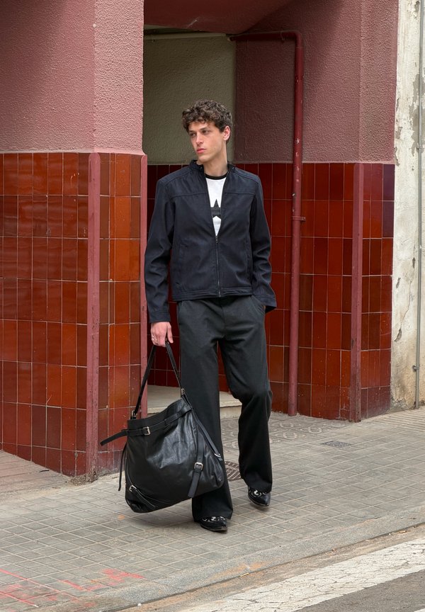 Νέος άνδρας με μαύρο μπουφάν και παντελόνι περπατάει στο πεζοδρόμιο, κρατώντας μια μεγάλη μαύρη δερμάτινη τσάντα κοντά στη γωνία ενός κτιρίου με κόκκινα πλακάκια.