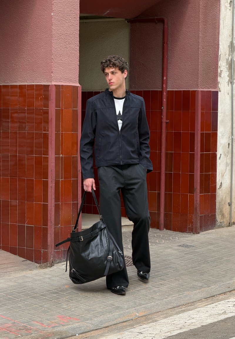 Jeune homme en veste et pantalon noirs marchant sur le trottoir, tenant un grand sac en cuir noir près d’un coin de bâtiment aux tuiles rouges.