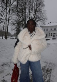 Femme portant un manteau en fausse fourrure blanc et un jean, tenant un sac à main rouge, debout sur un sol couvert de neige avec des arbres nus et une grande maison blanche en arrière-plan.