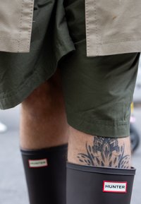 Botas de chuva de borracha castanhas com um logo "HUNTER" em branco e vermelho. Calções verde-oliva revelam uma tatuagem detalhada em preto na perna.
