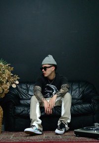 Čierna kožená pohovka, svetlosivá čiapka, čierne slnečné okuliare, tričko s potlačou, krémové nohavice s bočnými pruhmi, čierno-biele tenisky, nenápadná rastlina.