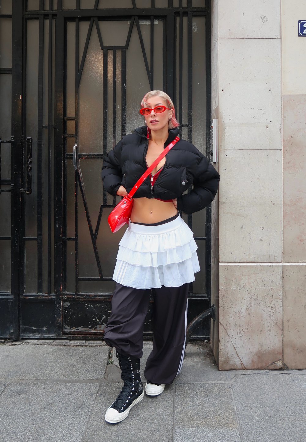 Personne aux cheveux blonds courts portant des lunettes de soleil rouges, une doudoune noire courte, une jupe blanche à volants par-dessus un pantalon noir, et des baskets montantes noires.