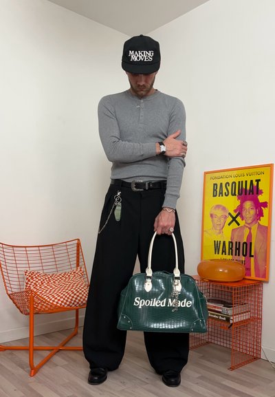 Hombre con camisa gris de manga larga y pantalones negros sosteniendo una bolsa verde de "Spoiled Made", lleva una gorra con la inscripción "Making Moves", de pie junto a una silla naranja y un cartel de Basquiat.