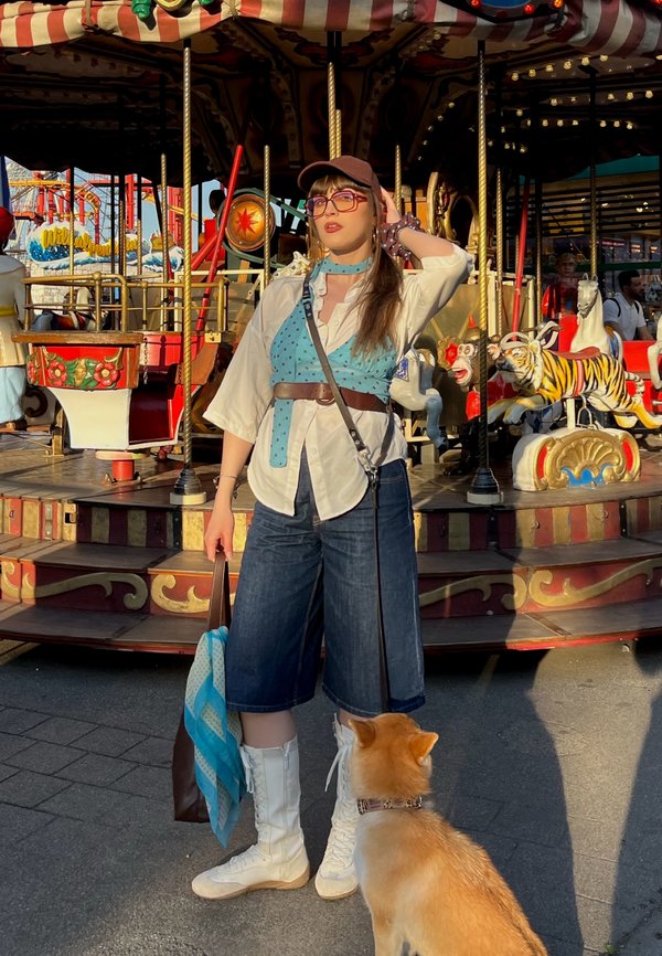 Kvinde i denimshorts og hvide støvler står med en lille hund foran en farverig karrusel i en forlystelsespark.