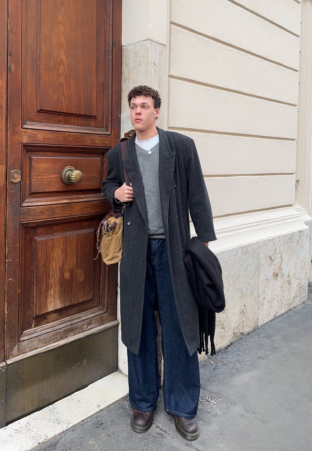 Jeune homme en long manteau sombre et large jean debout contre un mur en pierre près d'une grande porte en bois, tenant un sac et une écharpe pliée.