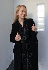 Abrigo de lana negro sobre una camisa negra a cuadros con líneas blancas, con cierre de botones en la parte delantera y mangas largas, sobre un fondo de pared clara.