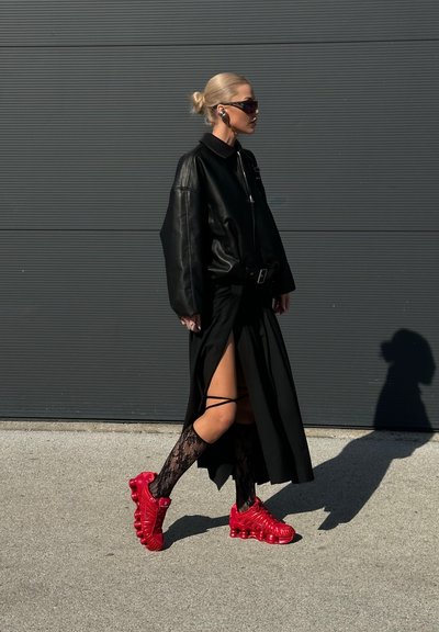 Mujer con el cabello rubio recogido en un moño camina vistiendo una chaqueta de cuero negra oversize, falda negra con abertura en el muslo, calcetas de encaje y zapatillas deportivas rojo brillante.