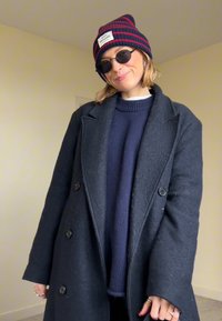 Femme souriante à l'intérieur portant des lunettes de soleil rondes, un bonnet rayé, un manteau sombre surdimensionné, un pull bleu marine et des bagues aux doigts.