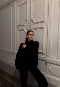 Pull à col roulé noir oversize en maille, tissu texturé ; le mannequin se tient contre un mur blanc à panneaux, tenant un sac, avec des cheveux lisses et des boucles d'oreilles.