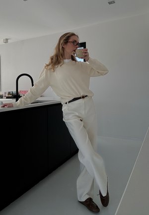 Persona con suéter crema y pantalones blancos apoyada en la isla negra de la cocina, bebiendo de una taza beige en una cocina moderna y minimalista.