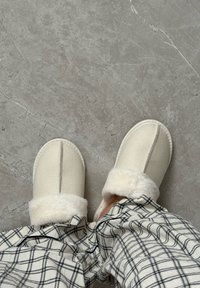 Pieds portant des chaussons pelucheux couleur crème et un pantalon de pyjama à carreaux, debout sur un sol en marbre gris.