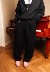 Personne portant un sweat à capuche noir et un pantalon, les mains dans les poches, debout à côté d'un piano en bois sur un sol en parquet, chaussée de chaussures roses.