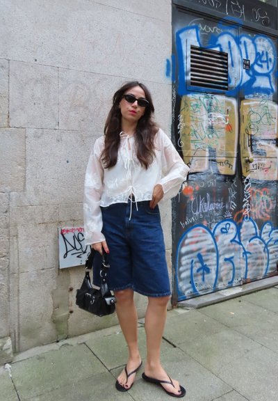 Mujer con cabello largo y oscuro que lleva gafas de sol, blusa blanca translúcida, pantalones cortos de mezclilla y chanclas, de pie sosteniendo un bolso negro junto a una pared cubierta de grafitis.