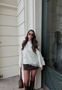 Femme aux longs cheveux bruns portant des lunettes de soleil, un pull blanc avec des nœuds, un haut en dentelle, une mini-jupe marron, des bottes montantes, tenant un sac à main marron près d'un bâtiment.