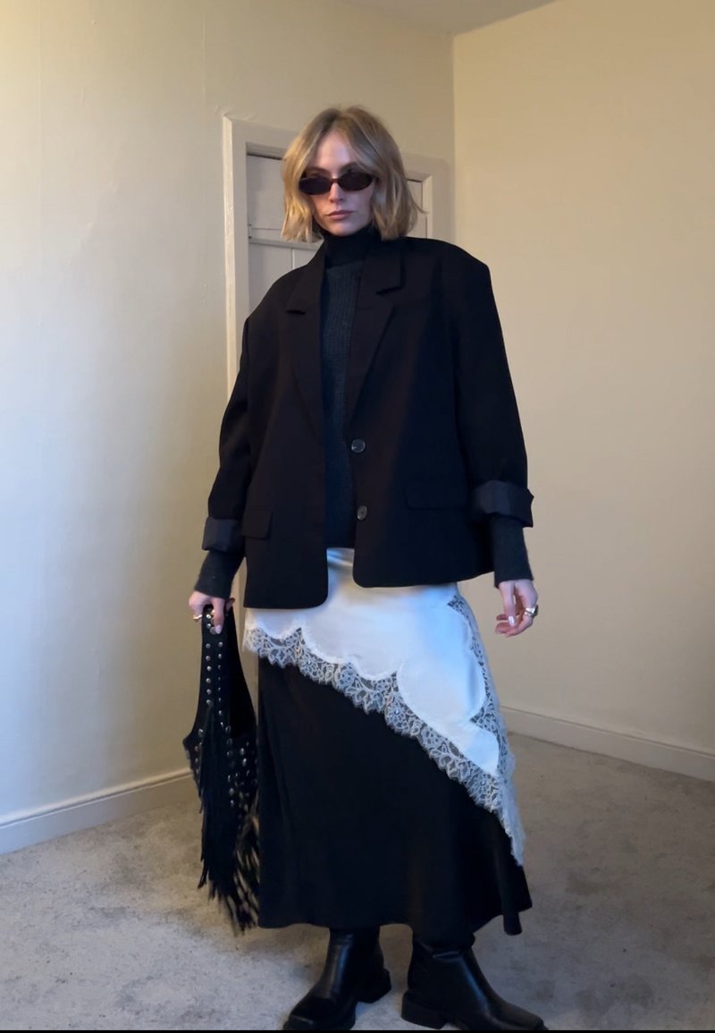Personne portant un blazer noir, un col roulé noir, des bottes noires, une longue jupe noire avec un détail en dentelle blanche, des lunettes de soleil noires, tenant un sac à franges noir clouté.