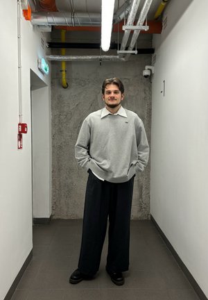 Sudadera gris con cuello y logotipo, combinada con pantalones negros holgados y zapatos negros. Pared de concreto y luces industriales en el fondo.