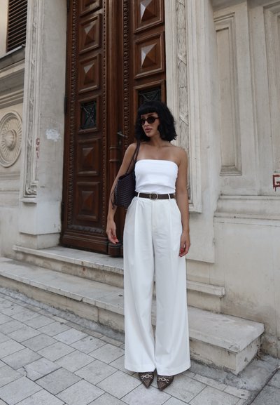 Top blanco sin tirantes, pantalones anchos de cintura alta blancos con un cinturón marrón, sosteniendo un bolso tejido oscuro, tacones puntiagudos marrones, de pie al aire libre.