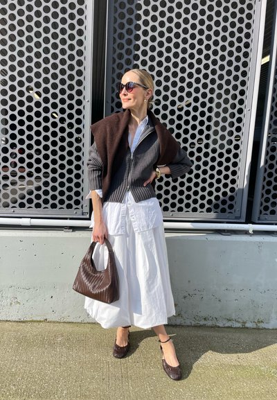 Mujer con gafas de sol, suéter gris con cremallera y chal marrón, falda larga blanca, sosteniendo un bolso marrón texturizado, de pie junto a una pared de metal perforado.