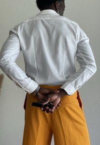 Person in a white long-sleeve shirt and mustard pants stands against a wall holding black sunglasses behind their back.
