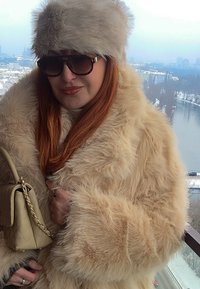 Femme portant un chapeau en fausse fourrure beige et un manteau, des lunettes de soleil, tenant un sac à main beige, debout sur un balcon donnant sur un paysage urbain enneigé au bord de la rivière.