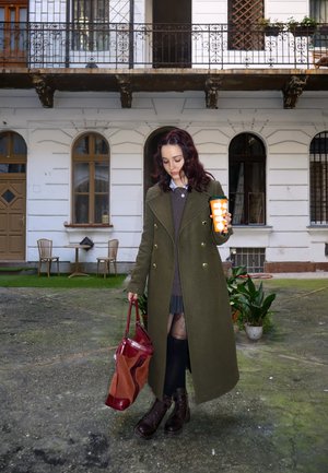 Abrigo largo en verde oliva con diseño de doble botonadura, suéter gris, falda plisada, sosteniendo una taza naranja; bolso rojo y botas marrones.