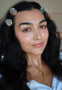 Una persona con lunghi capelli mossi e scuri adornati da fermagli a forma di conchiglia in tonalità pastello, che indossa una maglietta bianca e collane di oro a strati.