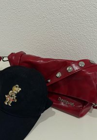 Čierna baseballová čiapka s vyšívaným medveďom v červenej sukni vedľa červenej kožené tašky Desigual so striebornými klincami na popruhu na bielom povrchu.