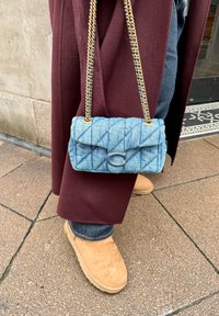 Sac bandoulière en denim matelassé avec une bandoulière chaîne dorée, équipé d'une fermeture à bouton métallique en forme de lettre "C". Porté avec un manteau bordeaux et des chaussures marron clair.