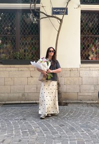 Žena v šedej tričku a krémovej sukni s čiernymi bodkami stojí na dlažobných kockách, drží kyticu kvetov a béžovú tašku.