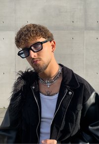 Czarna kurtka z korduroy z skórzanymi rękawami, nałożone srebrne łańcuszki, biały top na ramiączkach i czarne oprawki okularów. Kręcone włosy i widoczny tatuaż.