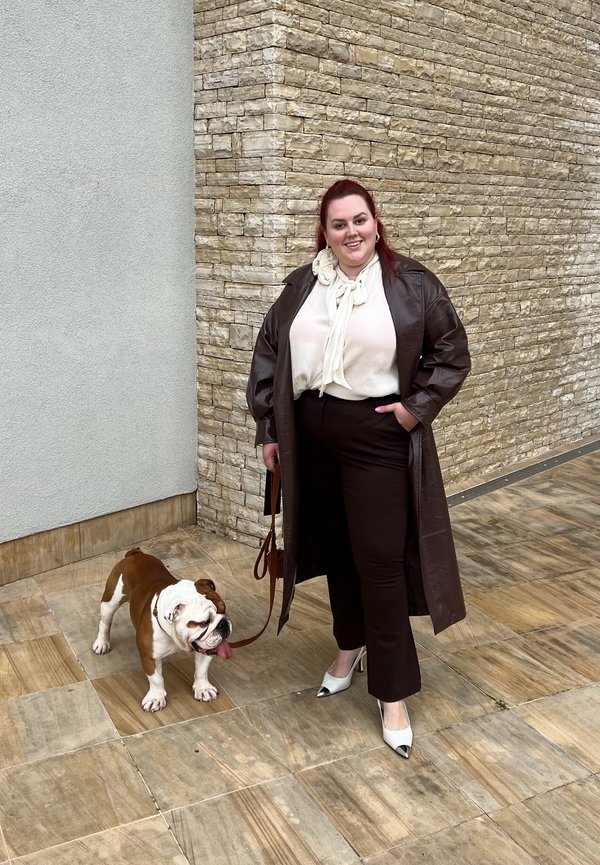 Hnědý kožený dlouhý kabát, krémová blůza s mašlí, tmavé kalhoty a špičaté bílé podpatky s černými akcenty. Bulldog na vodítku vedle osoby.