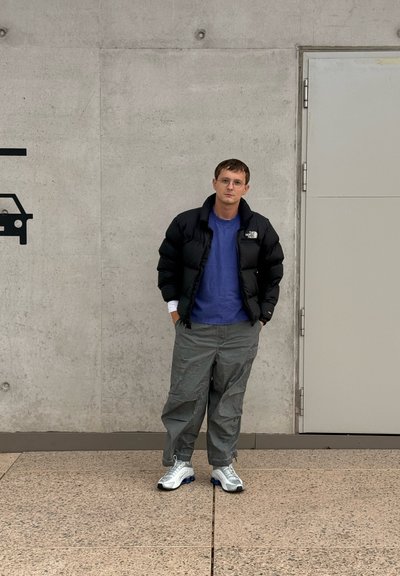 Chaqueta de plumas negra, camisa azul, pantalones de carga grises y zapatillas blancas con acentos azules. De pie contra una pared de hormigón gris.