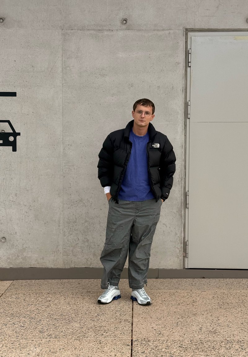 Zwarte donsjas, blauwe shirt, grijze cargobroek en witte sneakers met blauwe accenten. Staande tegen een grijze betonnen muur.