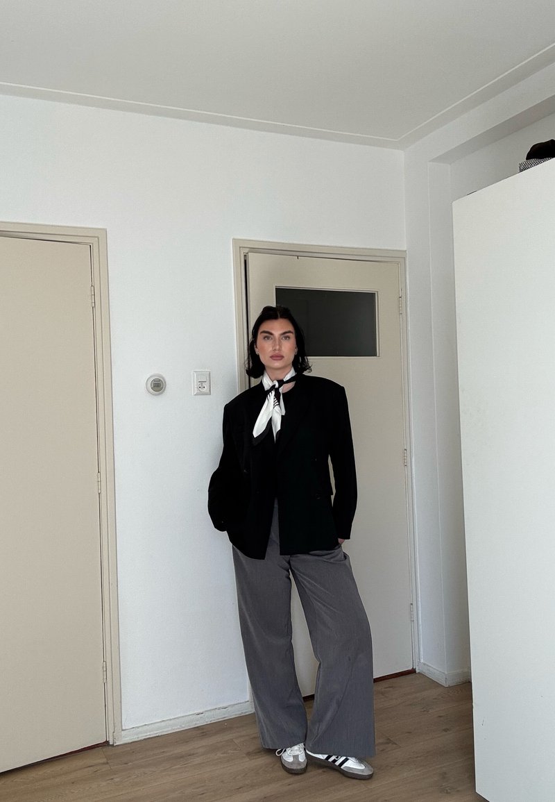 Personne aux cheveux foncés se tient à l'intérieur, portant un blazer noir, un pantalon gris à jambes larges, des baskets blanches et un foulard noir et blanc autour du cou, devant des murs blancs.