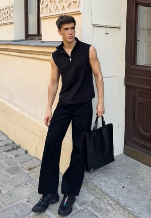 Zwarte mouwloze polo met rits, gecombineerd met wijde zwarte broek. Model draagt een matte zwarte tote bag en heeft zwarte sportschoenen aan.