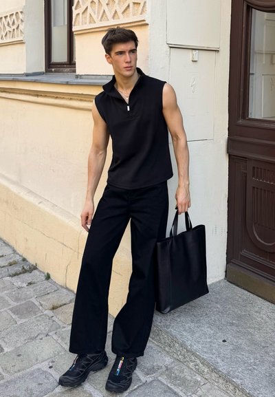 Polo negro sin mangas con cremallera, combinado con pantalones anchos negros. Modelo llevando un bolso tote negro mate, vistiendo zapatillas deportivas negras.