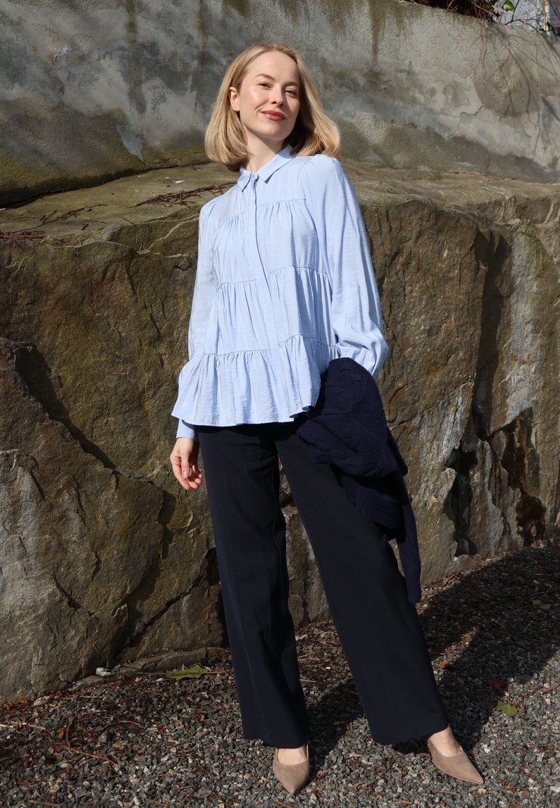 Femme blonde debout dehors contre un mur de pierre, portant un chemisier bleu clair à volants, un pantalon noir à jambes larges, des talons beiges, tenant un pull bleu marine.