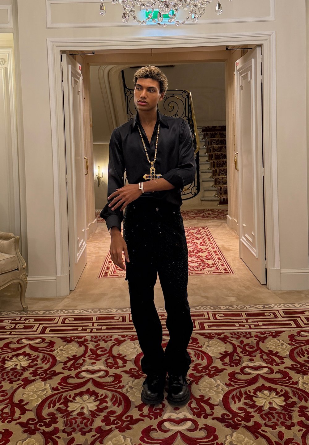 Jeune homme en tenue noire avec un grand collier croix, debout dans un couloir orné avec un tapis à motifs et un escalier derrière lui.